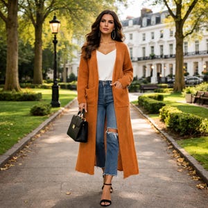 Op de afbeelding: Een vrouw draagt een lange oranje cardigan, een witte top en een gescheurde blauwe jeans, met een zwarte handtas. Ze draagt zwarte sandalen met hoge hakken. De achtergrond toont een park met bomen, een pad en gebouwen.