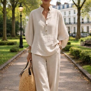 Puede incluir: Conjunto de lino beige compuesto por una parte superior de manga larga con cuello mandar&iacute;n y cierre de botones, combinado con pantalones de pernera ancha a juego. El conjunto se complementa con pulseras doradas y un bolso tote tejido. Ideal para el clima c&aacute;lido.