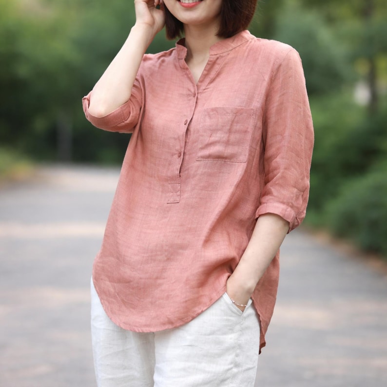 May include: A dusky pink linen shirt with a mandarin collar and a single chest pocket. The shirt features three-quarter sleeves and a relaxed fit. It's paired with white linen trousers, creating a casual, comfortable outfit.