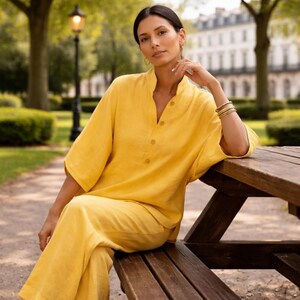 Puede incluir: Una persona con un conjunto de lino amarillo, que consta de una camisa con botones y pantalones a juego, sentada en un banco de madera en un parque. El conjunto tiene un corte holgado y un cuello mandar&iacute;n. Se ven joyas doradas, incluidos aretes y pulseras. El fondo muestra un parque con &aacute;rboles y edificios.