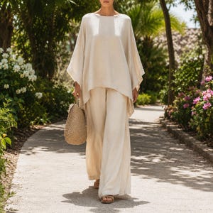 May include: A woman wearing a cream-colored linen outfit, including a loose-fitting top and wide-leg pants. She carries a woven straw handbag and wears sandals. The outfit is perfect for warm weather. The background features a stone path and lush greenery.