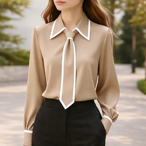 Può includere: Una camicetta beige con colletto e cravatta bordati di bianco, abbinata a pantaloni neri. La parte superiore a maniche lunghe presenta dettagli bianchi sui polsini. L'outfit è adatto per affari o occasioni formali.