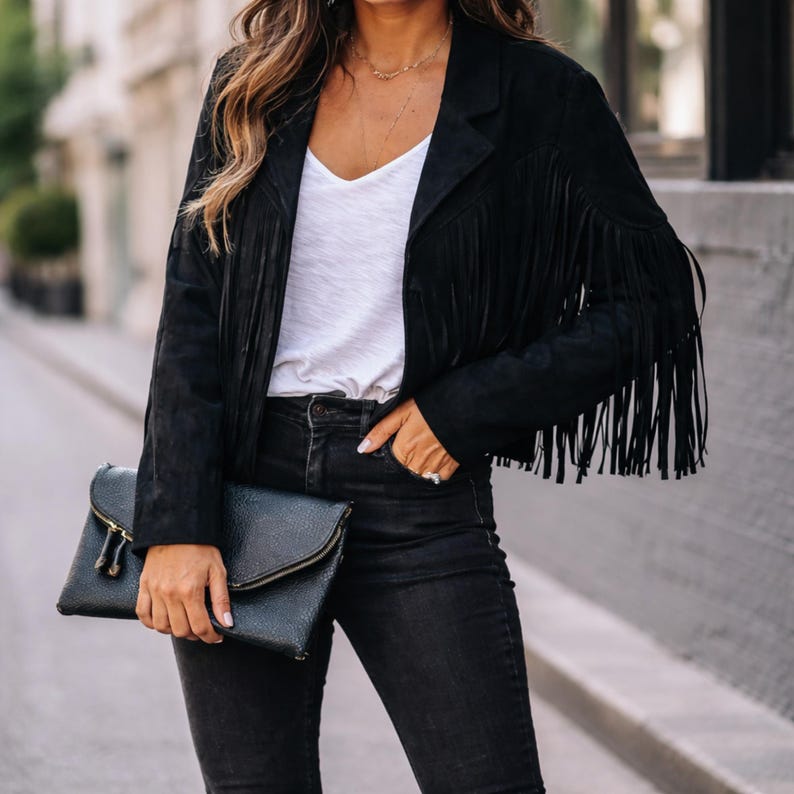 May include: Black suede fringe jacket with a white top and black jeans. A black clutch with a gold zipper is held in the hand. The jacket has a classic collar and long fringe along the sleeves and hem.