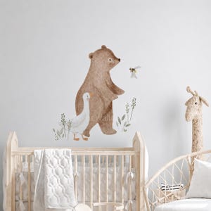 Può includere: Adesivo murale ad acquerello con un orso marrone, un'oca bianca e un'ape. L'orso è in piedi, con l'oca che cammina accanto. Il fogliame verde accentua la scena. Una testa di giraffa e una culla sono in primo piano.