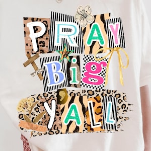 Puede incluir: Camiseta color crema con las palabras "PRAY BIG Y'ALL" en un diseño colorido y en capas. El diseño incluye estampado de leopardo, rayas y detalles florales. También se incluye una cruz y un lazo.