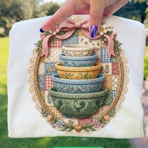 以下が含まれることがあります： 白いTシャツ。楕円形のフレームの中に、積み重ねられたカラフルなボウルの詳細なイラストが描かれています。デザインには、パッチワークの背景、ピンクのリボン、花のアクセントが含まれています。Tシャツの下部には「DOWNLOAD NOW」と書かれた緑色のボタンがあります。