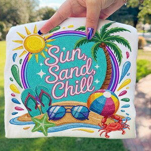May include: White t-shirt with a colorful beach-themed design. The design includes a sun, palm tree, sunglasses, flip-flops, beach ball, and a crab. The text "Sun, Sand, Chill" is also featured in the design.