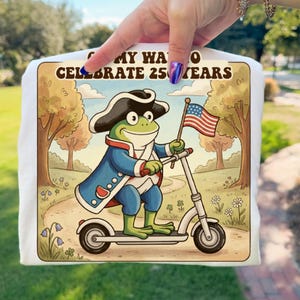 Könnte beinhalten: Weißes T-Shirt mit einem Cartoon-Frosch in einem Dreispitz und blauem Mantel, der einen Roller fährt und eine amerikanische Flagge hält. Das Bild hat den Text "My Way to Celebrate 25 Years". Der Hintergrund zeigt einen Weg und Bäume.
