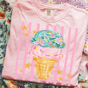 Retro pastel striped png featuring double scoop pastel ice cream cone top scoop has floral pattern and bottom scoop is pink.  Background stripes are pink yellow stars scattered.   Shown here on a folded blossom tee.