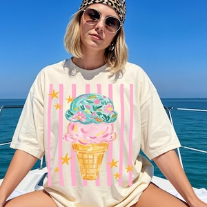 Retro pastel striped png featuring double scoop pastel ice cream cone top scoop has floral pattern and bottom scoop is pink.  Background stripes are pink yellow stars scattered.   Shown here on woman on a boat wearing a ivory oversized tee.   Ocean in the background.