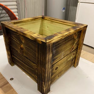 Peut inclure: Un bac à plantes carré en bois, teinté brun foncé et bordé d'un brun doré plus clair. Le bac a un design simple et rustique avec le grain du bois visible et est surélevé sur quatre pieds robustes. Pour l'intérieur ou l'extérieur.