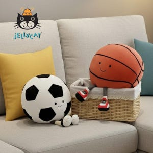 May include: A plush basketball and soccer ball with smiling faces sit on a couch next to a yellow pillow. The basketball has legs and red shoes, and is in a woven basket. The Jellycat logo is in the upper left corner.