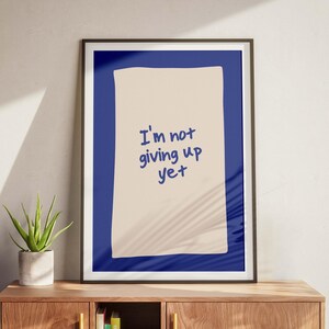 Könnte beinhalten: Gerahmter Kunstdruck mit dem Text "I'm not giving up yet" in blauer Schrift auf beigem Hintergrund, vor einem blauen Rand. Der Druck befindet sich in einem schwarzen Rahmen und wird auf einem Holzschrank mit einer Topfpflanze ausgestellt.