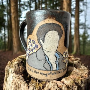 Könnte beinhalten: Eine Keramiktasse mit schwarzem Henkel und dunkelbrauner Außenseite. Die Tasse zeigt eine handgezeichnete Illustration einer Person im Anzug mit dem Text "This is the mug of a killer folk". Der Hintergrund ist hellbraun mit floralen Akzenten.