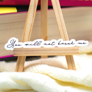 May include: A white sticker with a die-cut edge featuring the black cursive text "You will not break me" displayed on a small wooden easel. The sticker is positioned horizontally across the front support bar of the light-colored wooden stand.