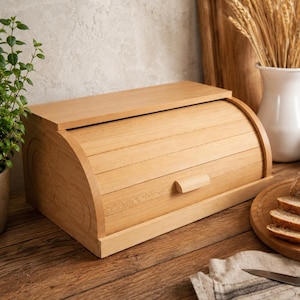Op de afbeelding: Een houten broodtrommel met een oprolbare deksel en een kleine houten handgreep. De broodtrommel heeft een gebogen voorkant. De broodtrommel staat op een houten oppervlak, met een brood en een mes in de buurt.