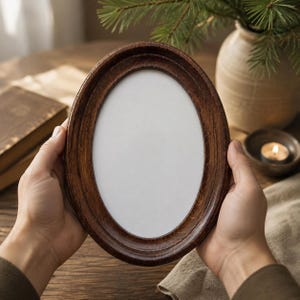 May include: An oval-shaped picture frame crafted from dark brown wood with a visible grain texture. The frame features a smooth, rounded profile and a wide border surrounding a blank, white, oval-shaped center area for displaying a photograph.