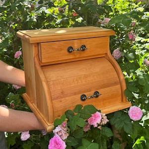 Peut inclure: Une boîte à pain en bois brun clair dotée d'une surface supérieure plate, d'un tiroir supérieur unique avec une petite poignée en métal et d'une porte roulante incurvée en dessous avec une poignée en métal assortie en bois naturel.