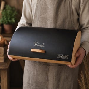 Puede incluir: Una panera de color negro mate con tapa curva y base de madera clara. La parte frontal presenta un pequeño tirador de madera, la palabra Bread en letra cursiva blanca y una etiqueta rectangular con el texto Maestro en fuente blanca.