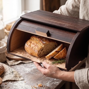 May include: A dark brown wooden bread box with a curved top and a pull-down door. Inside, a loaf of crusty bread is partially sliced, with a few slices visible. The bread box is held by a person, showcasing its design and functionality.