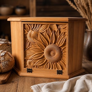 Peut inclure: Une boîte à pain en bois brun clair présentant une sculpture en relief détaillée de tournesols sur le panneau avant. La boîte a une forme rectangulaire avec une porte battante en bas, fixée par deux petites charnières en métal noir.