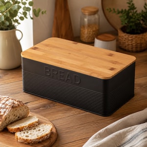 May include: A rectangular black metal bread bin with a horizontal ribbed texture and the word BREAD embossed on the front. The container features a flat, light-toned bamboo wood lid that sits securely on top of the matte black metal base.