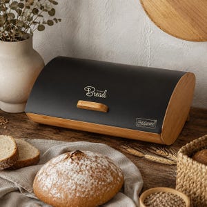 May include: A matte black bread box with a curved lid, featuring a light wood base and handle. The front displays the white text "Bread" in a script font and a small rectangular logo with the word "Maestro" in a decorative script typeface.