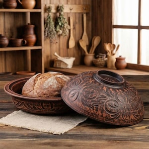 May include: A brown ceramic bread baker with a loaf of bread inside. The lid, decorated with floral patterns, is placed beside the baker. The scene is set on a wooden table, with a wooden background and kitchen utensils.