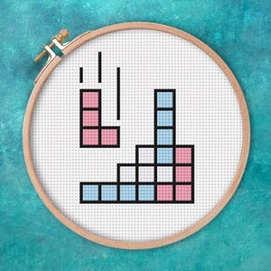 May include: A cross-stitch embroidery hoop with a Tetris game design. The design uses pink and light blue blocks with black outlines, set against a white background. The wooden hoop is secured with a brass clasp.