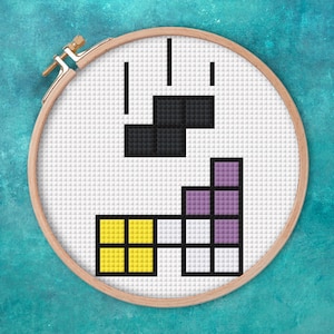 Può includere: Un telaio da ricamo a punto croce con un design del gioco Tetris. Il design include blocchi neri, gialli, bianchi e viola. Il telaio in legno è fissato con un fermaglio in ottone. Lo sfondo è una griglia bianca.