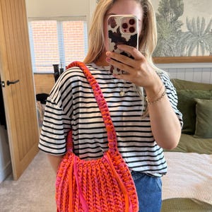 May include: A vibrant, handmade shoulder bag in shades of orange and pink. The bag features a braided strap and fringe details, adding texture and visual interest. The bag is a bold accessory.