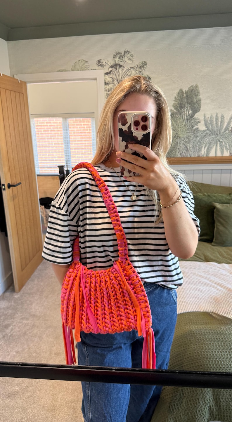 May include: A vibrant, handmade shoulder bag in shades of orange and pink. The bag features a braided strap and fringe details, adding texture and visual interest. The bag is a bold accessory.