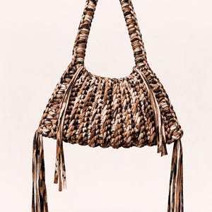 May include: A handcrafted, woven handbag in shades of brown, beige, and black. The bag features a braided handle and decorative fringe. The bag's texture and color palette suggest a natural, bohemian style.