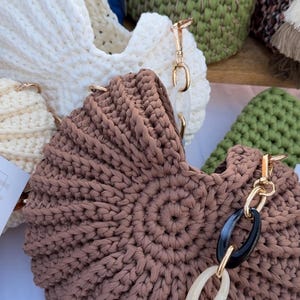 May include: A selection of handmade crochet bags in different colours. A brown bag with a gold chain and black and cream accents is in focus. Other bags are white and green. The bags are made of thick yarn.