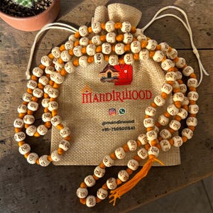 Peut inclure: Un chapelet de prière avec des perles en bois blanches et un fil orange, présenté avec un sac en toile de jute. Les perles portent du texte. Le sac porte le texte "MANDIRLIOOD" et des identifiants de réseaux sociaux.