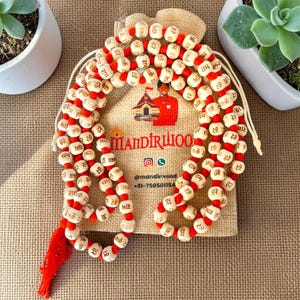 Puede incluir: Un collar de cuentas de oración con cuentas de madera clara y texto en sánscrito en rojo, ensartadas en un cordón naranja brillante con una borla a juego. El collar se presenta sobre una bolsa de arpillera beige con el texto Mandirwood.