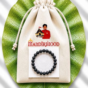 May include: A black lava stone bead bracelet on a white card with a geometric design, alongside a drawstring bag. The bag has a logo with text and an illustration of a person and a building. The items are displayed on a large green leaf.