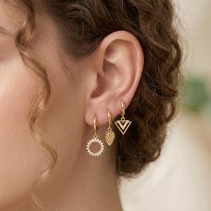 May include: Gold hoop earrings featuring three distinct pendant designs: a sunburst, a teardrop, and a triangle with a chevron pattern. The earrings are displayed on an ear, highlighting the jewellery's details and gleam.
