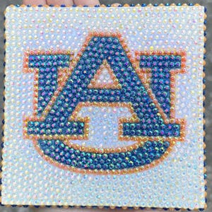 May include: A square decorative plaque covered in iridescent white rhinestones featuring the Auburn University AU logo in the center. The logo is outlined in orange rhinestones and filled with dark blue rhinestones, creating a textured surface.