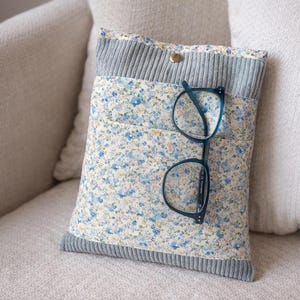 Könnte beinhalten: Eine Bucheinband mit floralem Muster, einem blauen Cord-Oberteil und einem goldenen Druckknopfverschluss. Eine Brille mit tealfarbenem Rahmen liegt auf der Vorderseite. Die Hülle dient zum Schutz von Büchern.