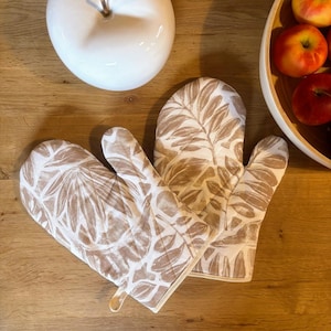 Könnte beinhalten: Zwei Ofenhandschuhe mit einem beige-weißen Blättermuster. Sie liegen auf einer Holzoberfläche, neben einem weißen Keramikapfel und einer Holzschale mit roten Äpfeln. Die Handschuhe haben eine Schlaufe zum Aufhängen.