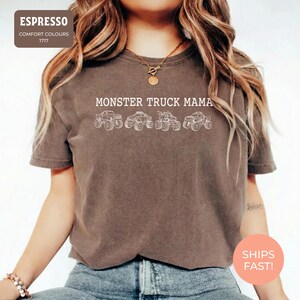 Espresso Comfort Colors t-shirt featuring the text "MONSTER TRUCK MAMA" and four white monster truck designs. The shirt has a crew neck and short sleeves.
