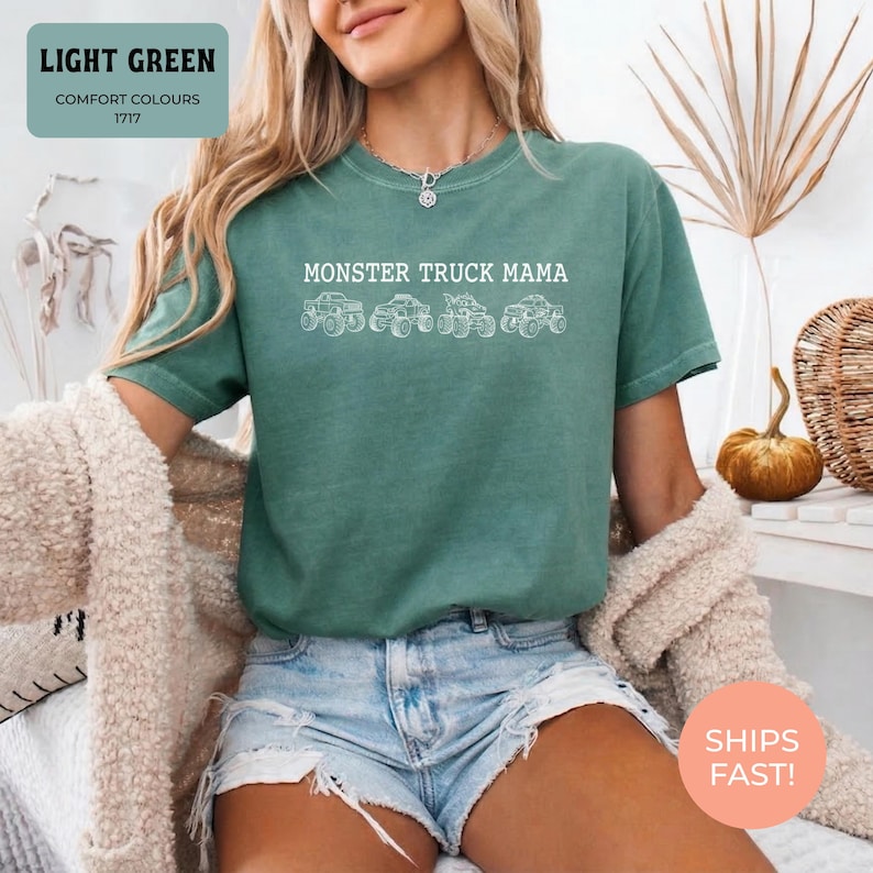 Light Green Comfort Colors t-shirt featuring the text "MONSTER TRUCK MAMA" and four white monster truck designs. The shirt has a crew neck and short sleeves.