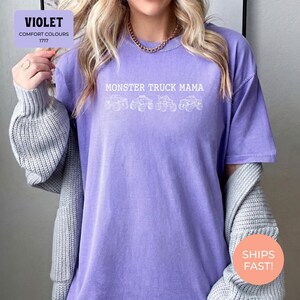 Violet Comfort Colors t-shirt featuring the text "MONSTER TRUCK MAMA" and four white monster truck designs. The shirt has a crew neck and short sleeves.
