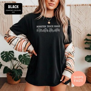 Black Comfort Colors t-shirt featuring the text "MONSTER TRUCK MAMA" and four white monster truck designs. The shirt has a crew neck and short sleeves.