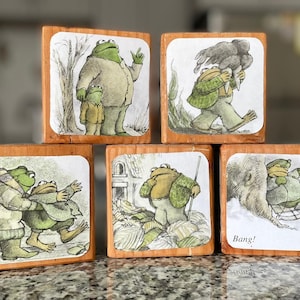 May include: Five square wooden blocks featuring illustrations of green frogs in various scenes. The images depict frogs walking, carrying items, and interacting, with one block showing the text Bang! printed in the lower left corner of the illustration.