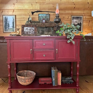 May include: A deep red wooden sideboard featuring two side cabinets with doors, two central stacked drawers with round knobs, and a lower open shelf. The piece stands on four turned legs and displays a visible wood grain texture throughout.