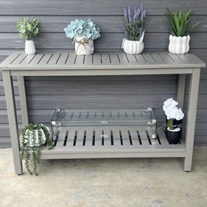 May include: A gray rectangular console table with a slatted top and lower shelf. The top holds four potted plants, including greenery, blue hydrangeas, lavender, and an aloe plant. The lower shelf features a glass fireplace and a trailing plant.