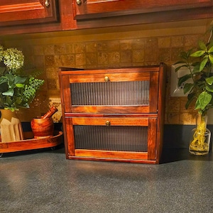 May include: A two-tiered wooden cabinet with a warm, reddish-brown finish. Each level has a door with a ribbed, semi-transparent panel and a small, round brass knob. The cabinet is designed for storage and organization.