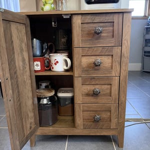 May include: A wooden coffee cabinet with four drawers and a cabinet door. The drawers have decorative floral knobs. The cabinet door is open, revealing mugs, a jar of coffee, and a container. A coffee maker sits on top.